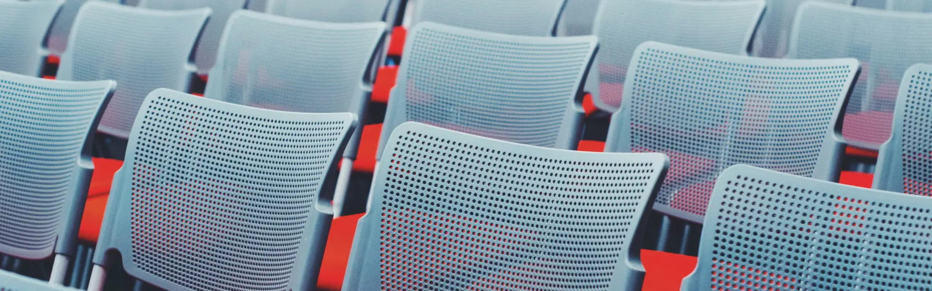 A pattern of empty, modern plastic chairs with perforated backs and red seats arranged in rows, viewed at an angle. The image creates a geometric, abstract visual effect with repetition and contrast between the cool-toned backs and vibrant red seats.